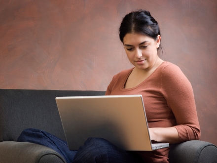 Women using a laptop computer. She is sitting on a couch looking at her computer screen. 